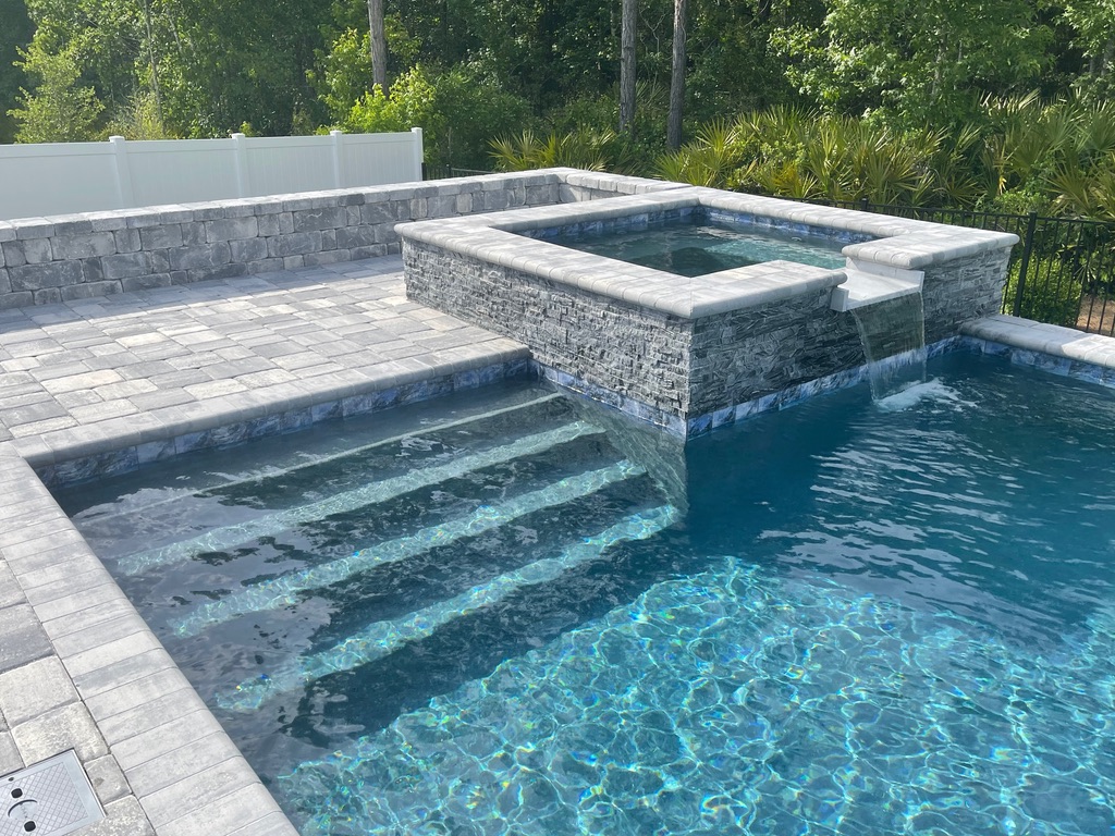 Custom pool design featuring a stone waterfall and spa, surrounded by paver patio and lush greenery, emphasizing Breakwater Construction's pool financing options.